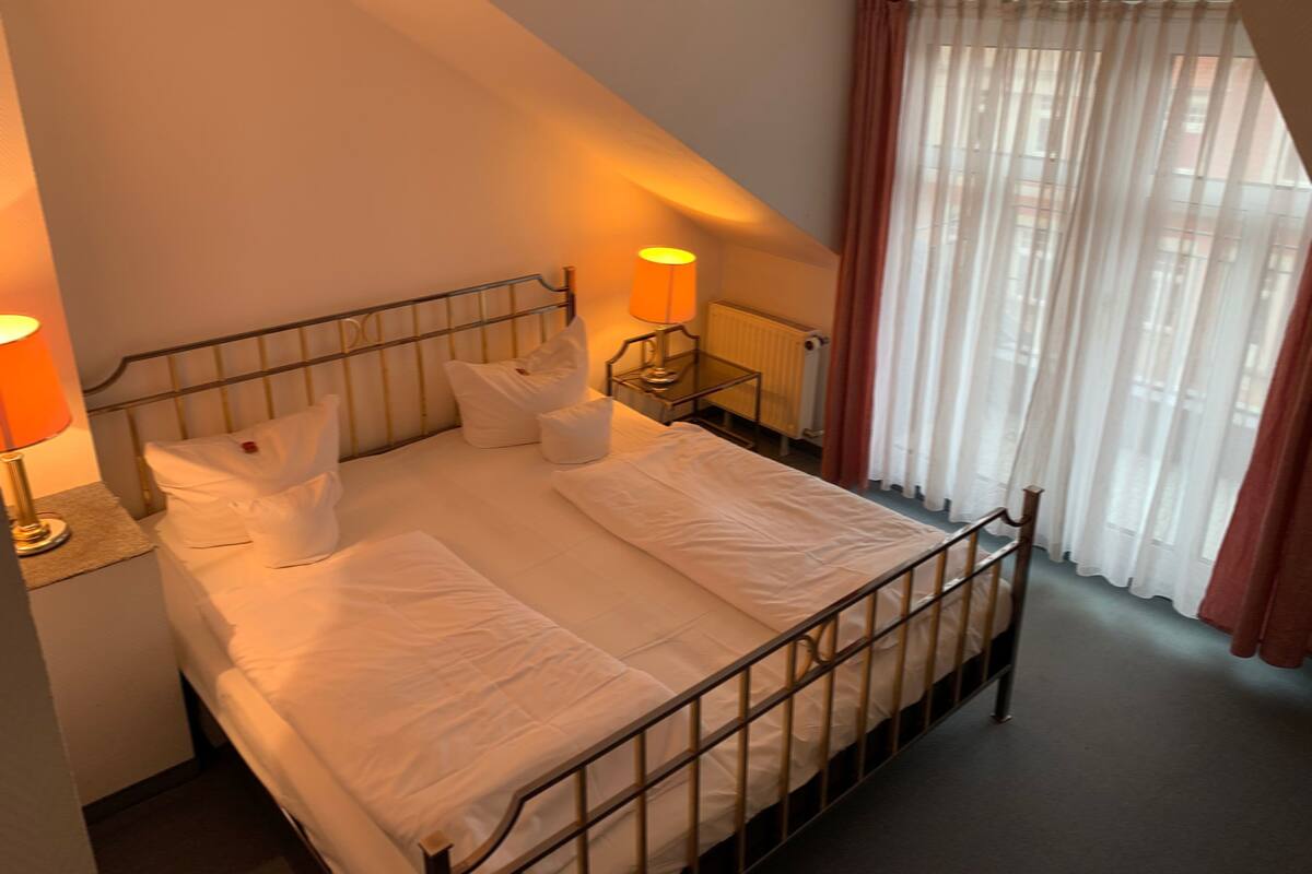 A neatly made bed in a room with a sloped ceiling, a bedside table with a lamp, and a window with sheer curtains.