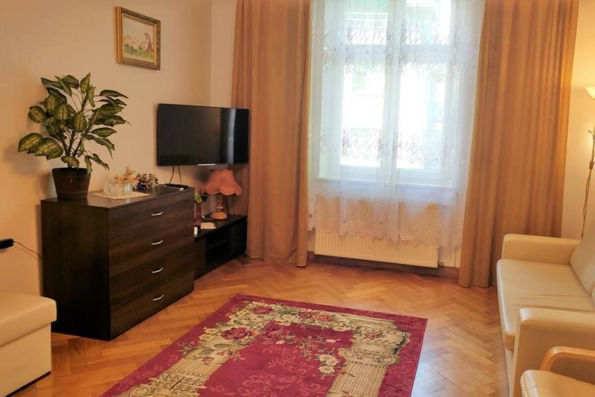 Un salón con un aparador de madera oscura, un televisor, un sofá beige, un tapete estampado y una ventana con cortinas traslúcidas.