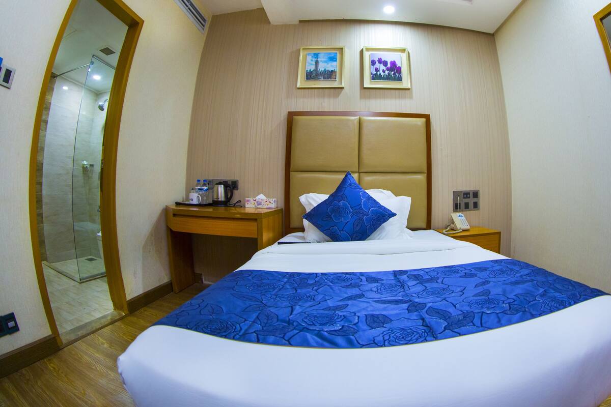 A hotel room with a large bed, a bedside table with a phone and a kettle, a mirror, and two framed pictures on the wall.