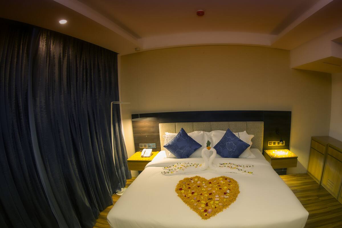 A hotel room with a large bed, a heart-shaped decoration on the bed, and a nightstand with a phone and lamp.