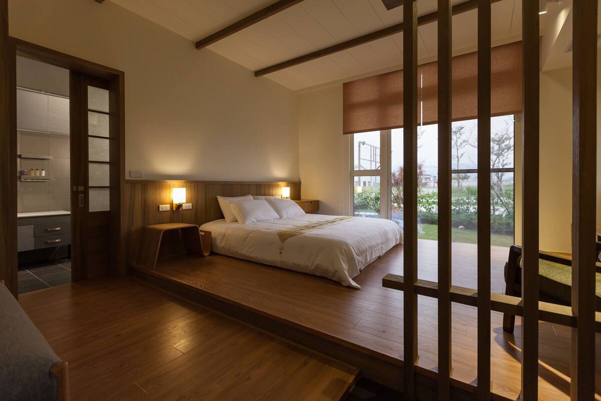 A bedroom with a wooden floor, a bed with white bedding, two bedside tables with lamps, and a large window offering a view of the outdoors.