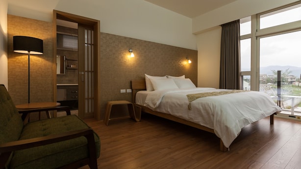 Una habitación de hotel con piso de madera, una cama con ropa blanca, un sillón verde, una mesita y una lámpara.