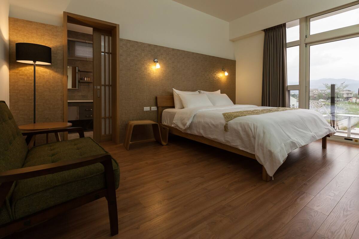 A hotel room with a wooden floor, a bed with white linens, a green armchair, a small table, and a lamp.