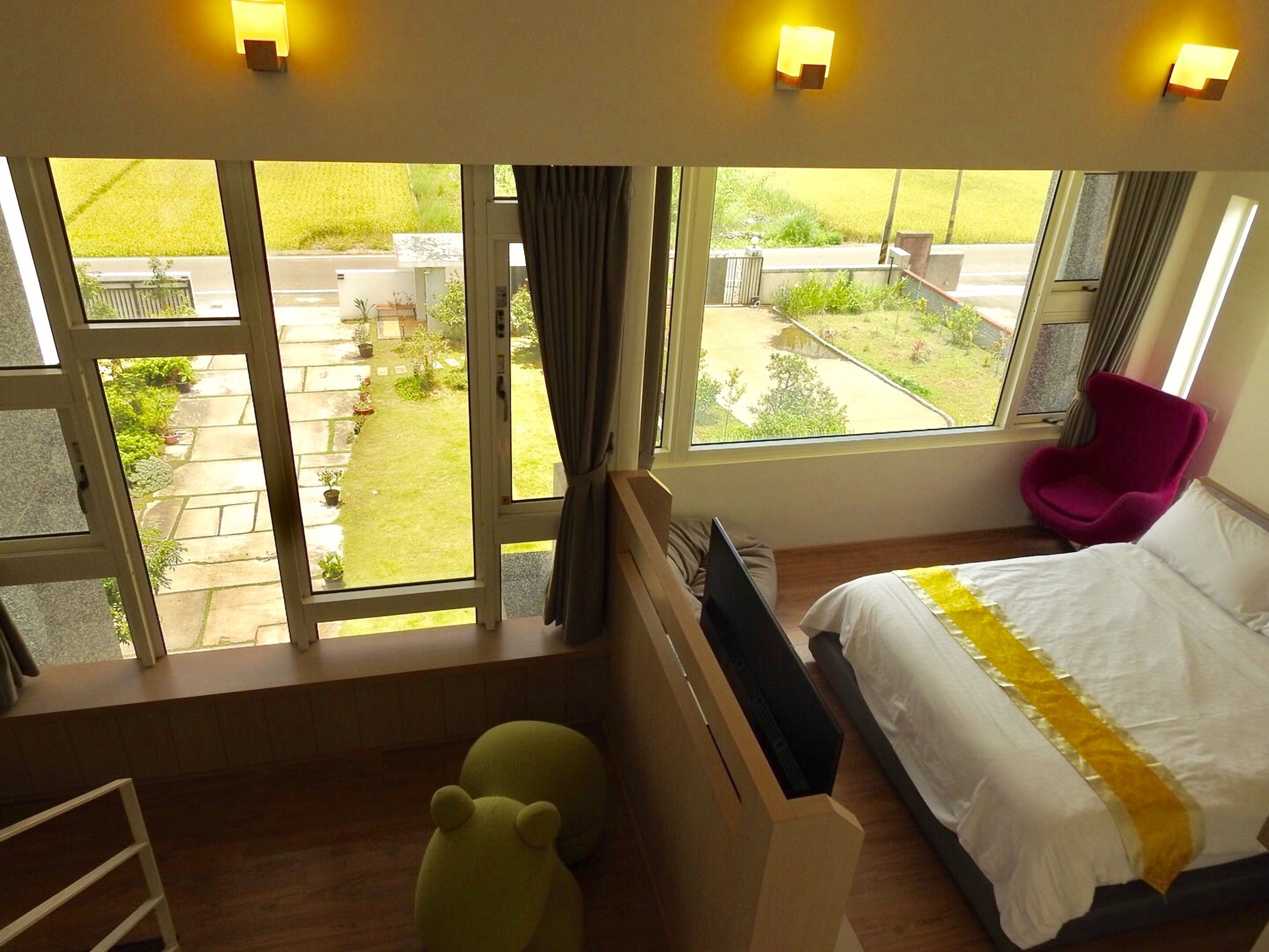 A hotel room with a bed, a desk, a chair, and a view of a garden through large windows.