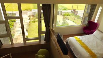 A hotel room with a bed, a desk, a chair, and a view of a garden through large windows.