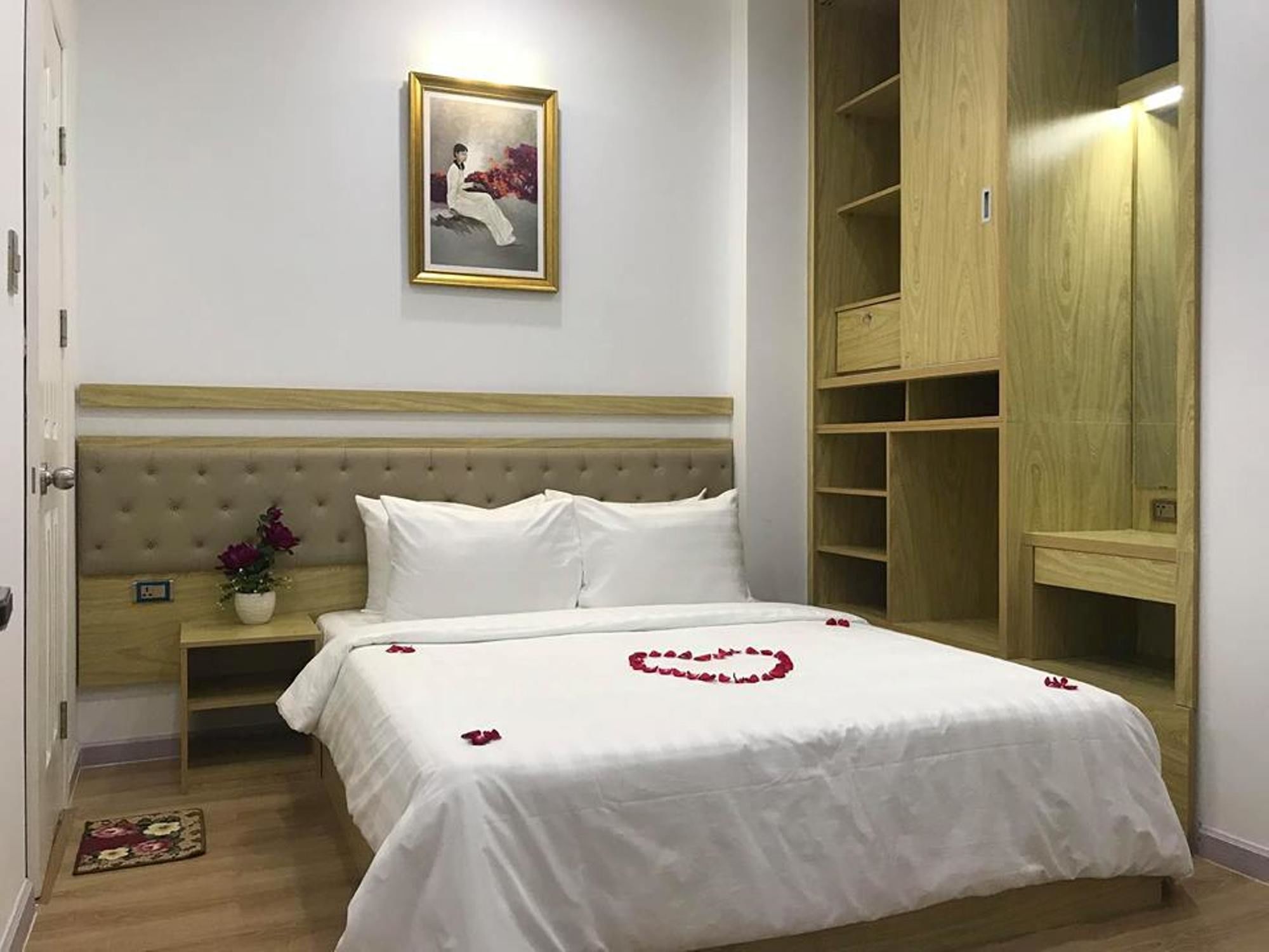 A bedroom with a bed, a nightstand, a bedside table with a vase of flowers, a wooden wardrobe, and a framed picture on the wall.