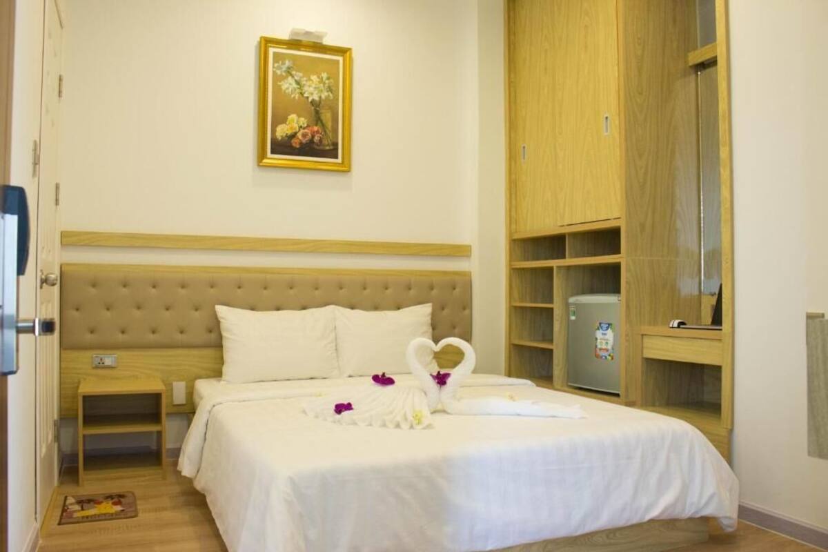 A hotel room with a large bed, a wooden headboard, a small bedside table, a nightstand, a wardrobe, and a framed picture on the wall.