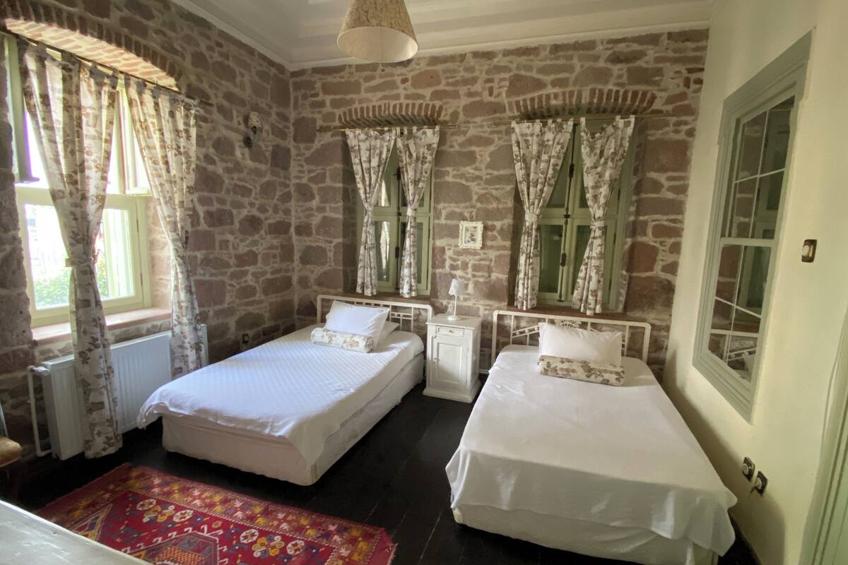 A room with two single beds, a small bedside table with a lamp, a red patterned rug, stone walls, and a window with curtains.