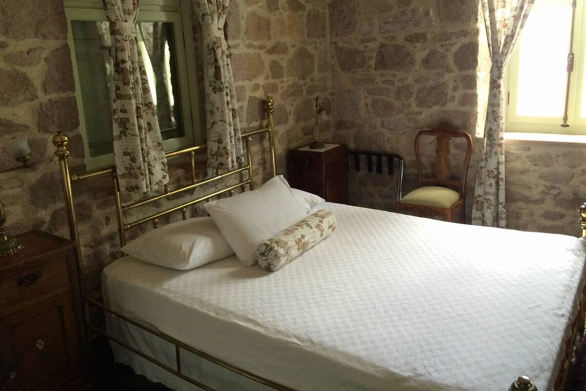A bedroom with a stone wall, a bed with a white bedspread, a nightstand, a chair, and a window with curtains.