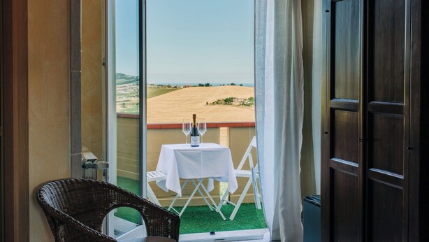 A room with a view of a field, a table with a bottle and glasses, and a wicker chair.