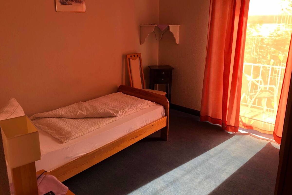 A single bed with a wooden frame, a small bedside table, a mirror, and a window with curtains.