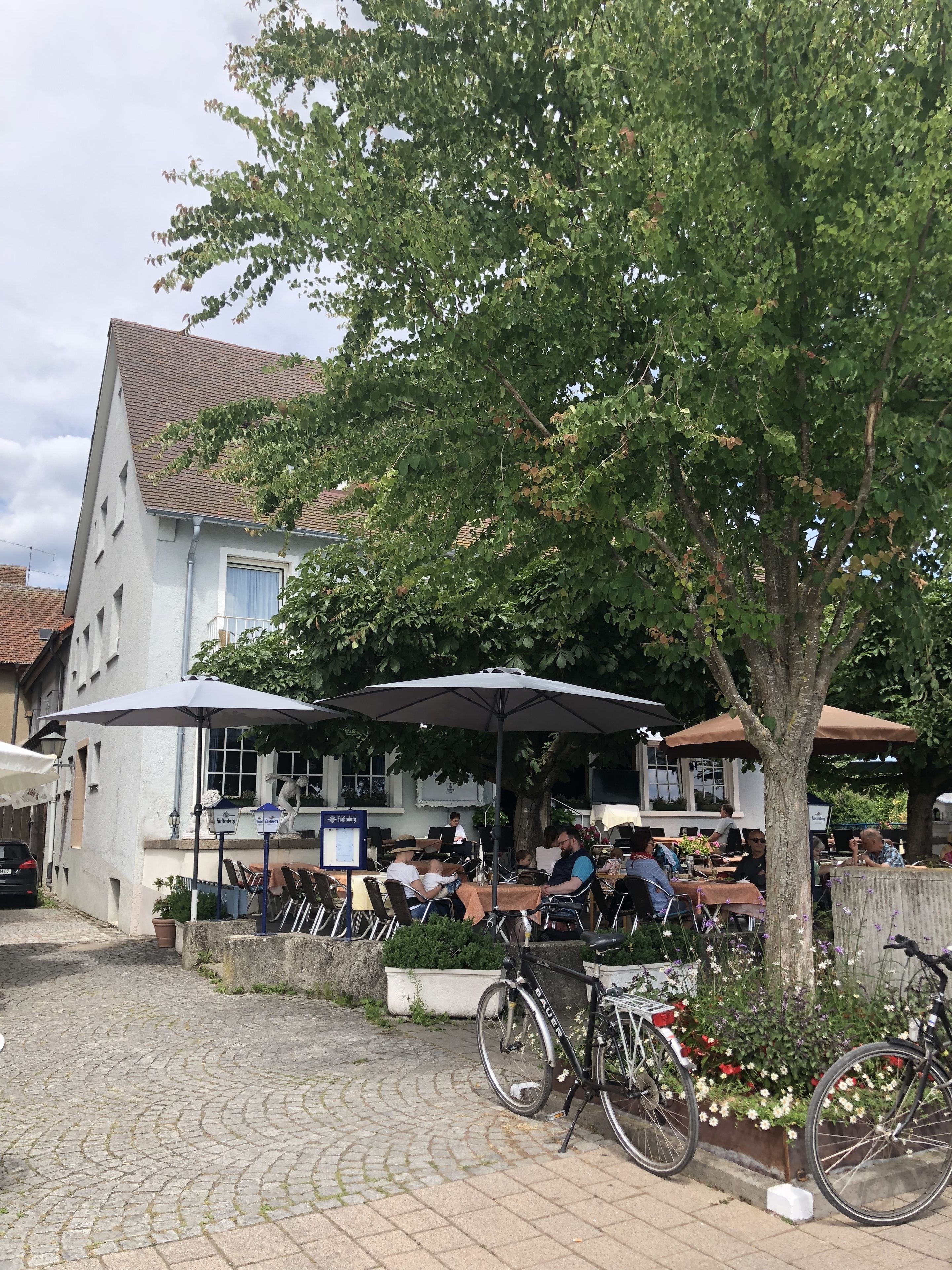 Ein Straßencafé mit Tischen und Stühlen, ein Fahrrad und im Hintergrund ein Gebäude mit Ziegeldach.