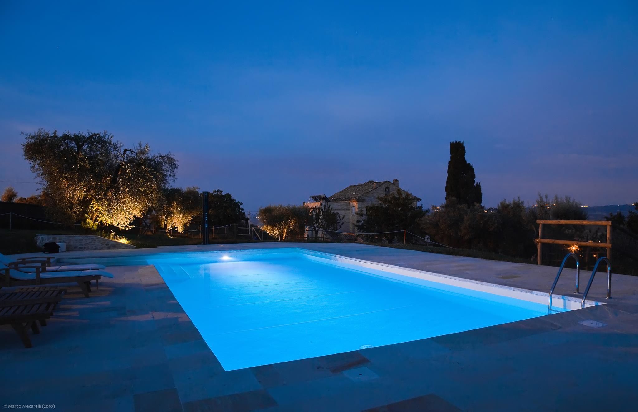 Une piscine dont le bord est illuminé de lumière bleue, entourée de chaises longues et d’un mur en pierre.
