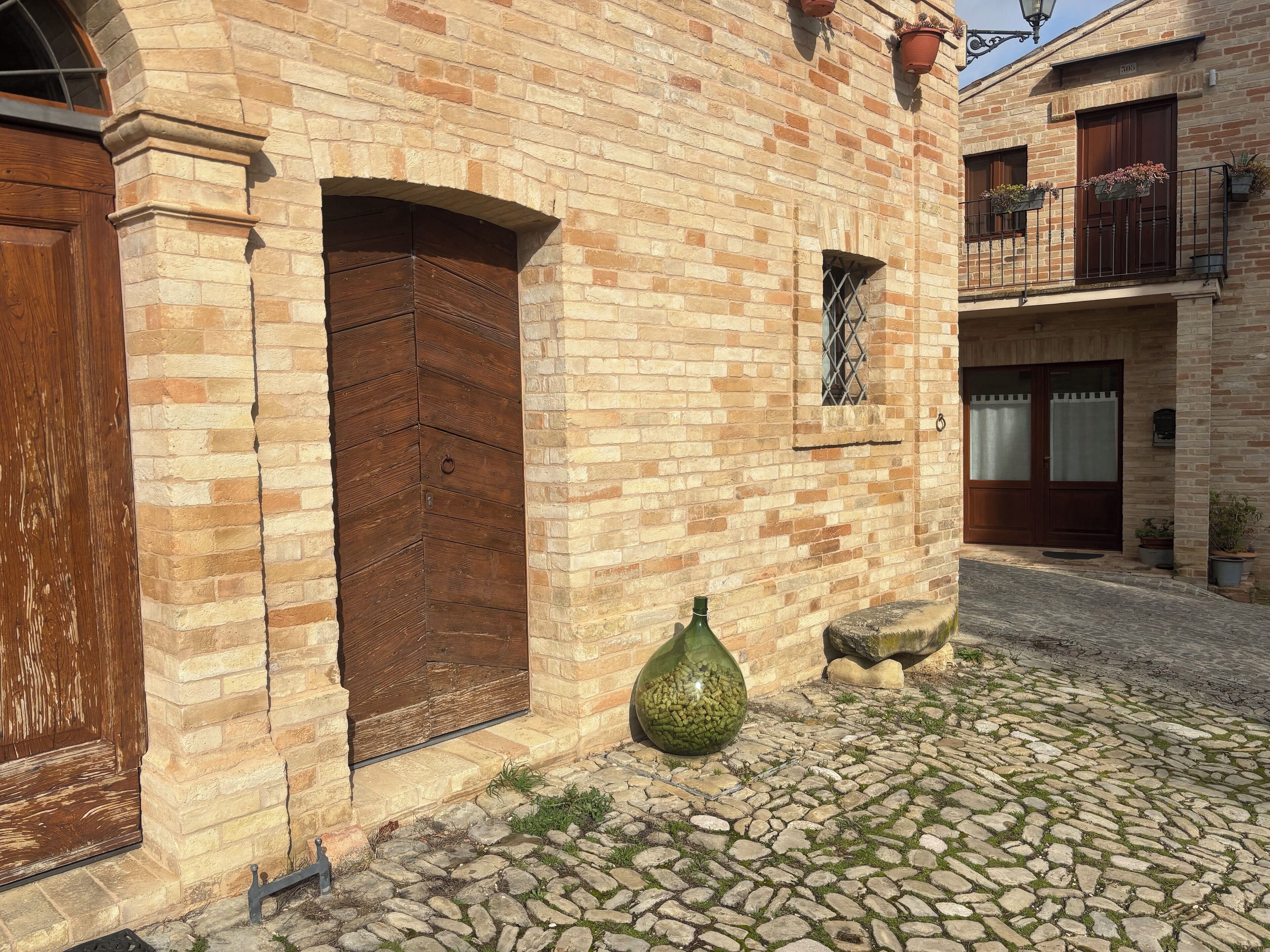 Un bâtiment traditionnel en briques, doté d’une grande porte en bois, d’une petite fenêtre ornée d’une grille métallique et d’une cour pavée.