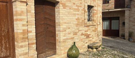 A traditional brick building with a large wooden door, a small window with a metal grille, and a cobblestone courtyard.