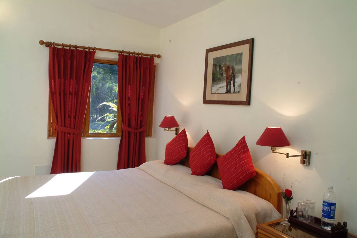 Een opgemaakt bed met rode kussens, een nachtkastje met een lamp, een ingelijst beeld aan de muur, en een raam met rode gordijnen.
