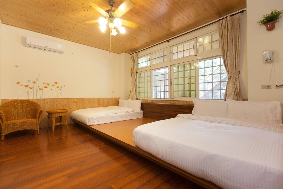 A room with two single beds, a wooden ceiling, a ceiling fan, a window with curtains, a wicker chair, and a small table.