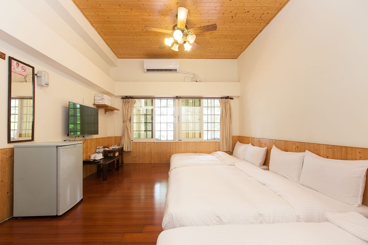 A hotel room with a wooden ceiling, a bed with white linens, a desk with a chair, a television, and a window with curtains.