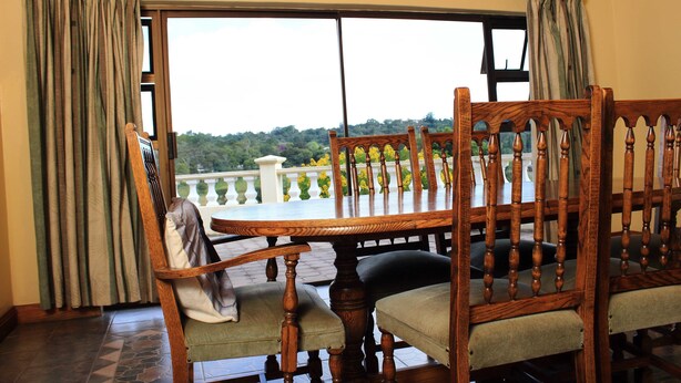 A dining area with a wooden table and chairs, a view of greenery through a large window, and a balcony with a railing. - Unique Bed and breakfast (Harare)
