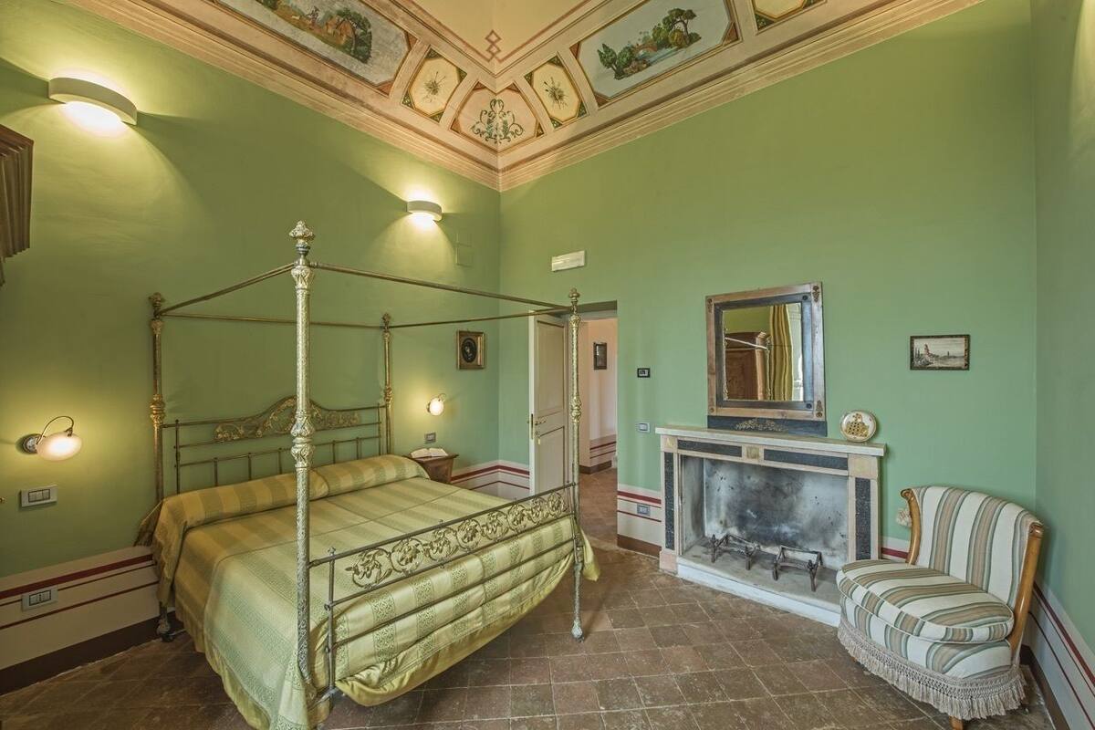 Una cama con dosel de estructura metálica y diseño ornamentado, una chimenea con repisa, y un sillón a rayas.
