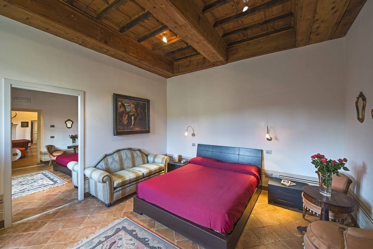 Un dormitorio con una cama grande, un sofá, una mesita y una silla. Hay un cuadro en la pared y una alfombra en el suelo.