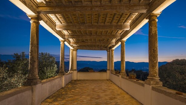 Un espacio exterior cubierto con columnas de piedra y suelo de baldosas, con vistas a un paisaje montañoso.