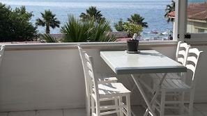 Ein Balkon mit weißem Tisch und Stühlen, einem Blumentopf und Blick auf das Meer und Palmen.