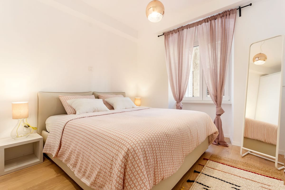 A bedroom with a bed, bedside lamps, a mirror, and a window with curtains.