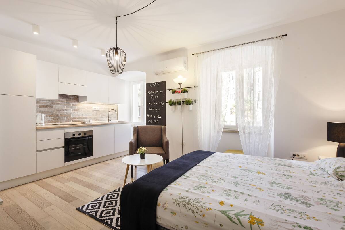 A modern hotel room with a kitchen, a bed with floral bedding, a small table with a plant, and a view of the outdoors.