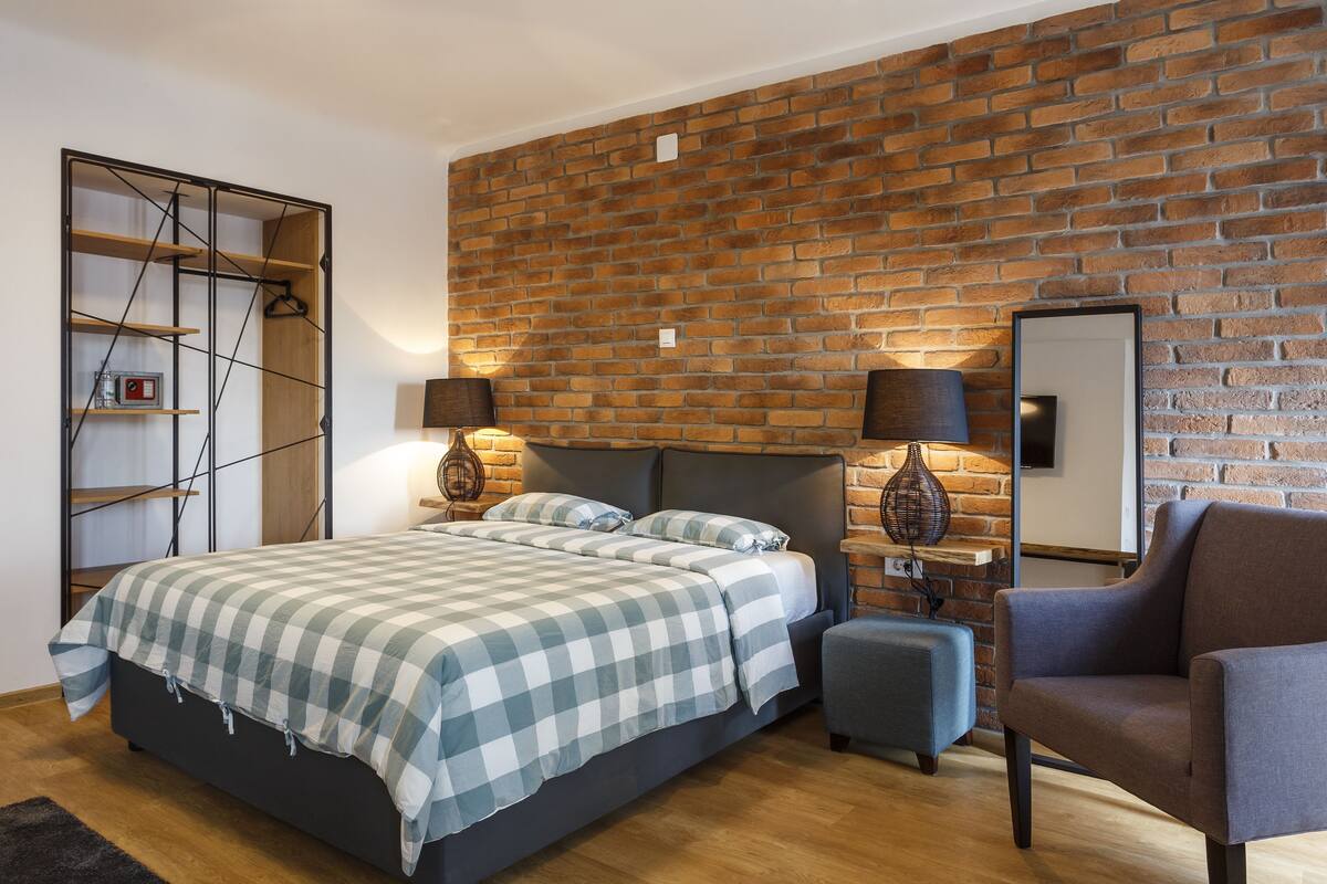 A bedroom with a brick wall, a bed with a checkered bedspread, two bedside lamps, a chair, and a hanging light fixture.