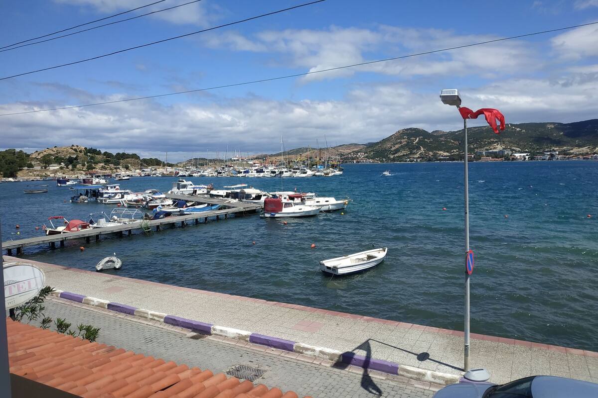 A marina with boats docked, a pier, and a view of the sea and hills in the background.