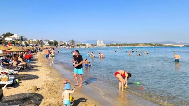 A beach with people swimming and sunbathing, a clear sky, and buildings in the background. - Hotel Delta Altinkum (Didim)