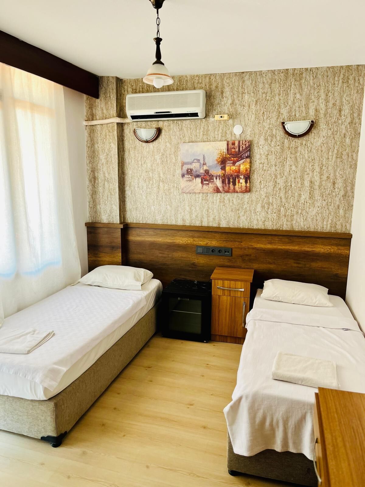 A hotel room with two single beds, a wooden headboard, an air conditioning unit, a painting on the wall, and a small wooden cabinet.