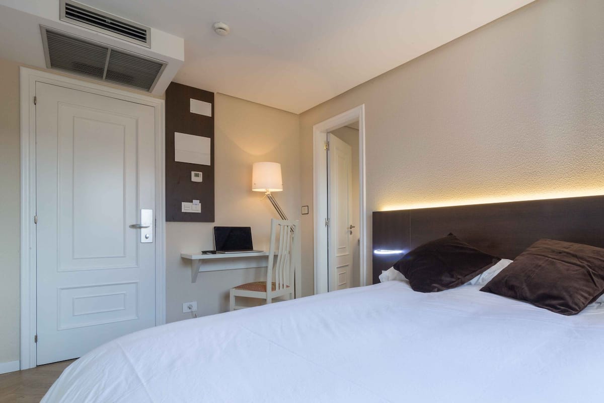 A hotel room with a bed, a desk with a laptop, a chair, a lamp, and a door.
