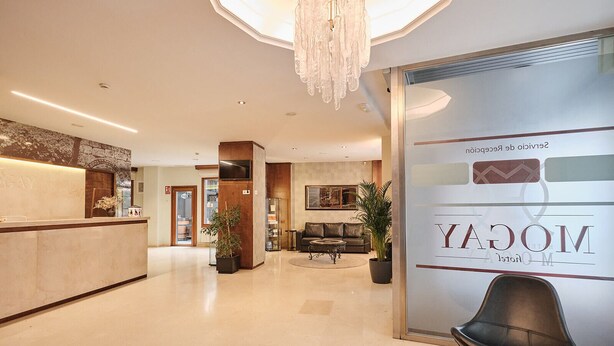 A hotel lobby with a reception desk, seating area, and a large window with the hotel name. - Hotel Mogay (Chantada)