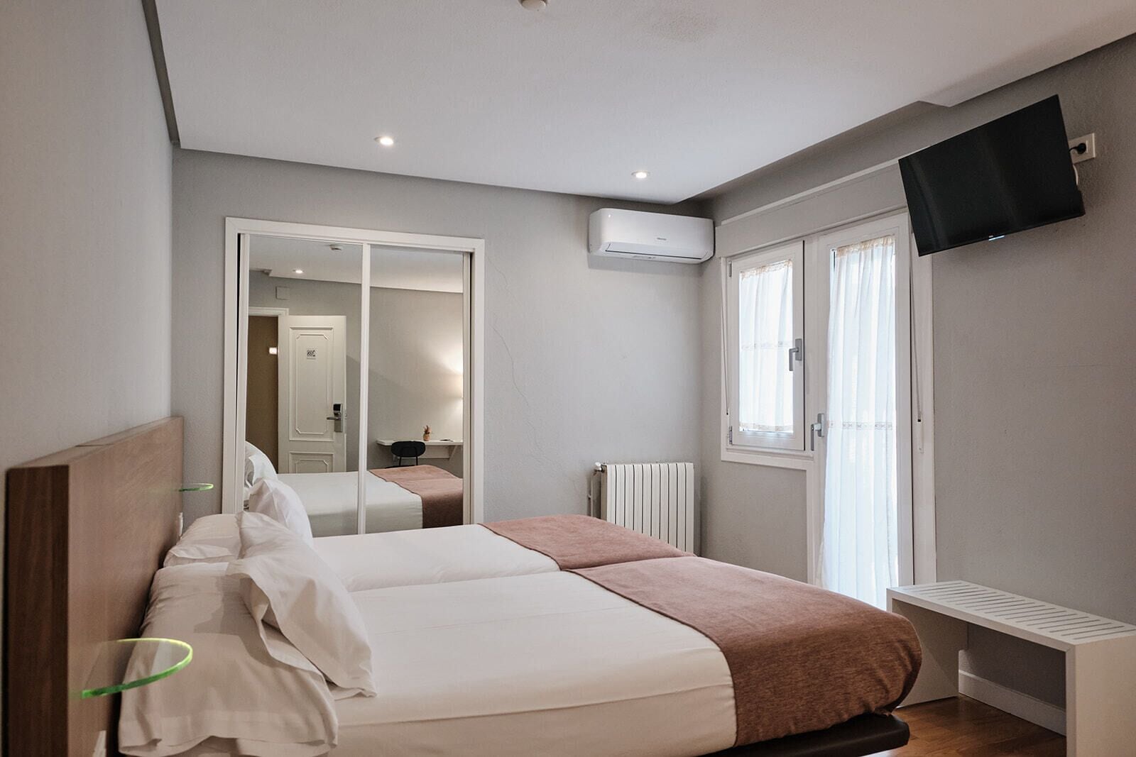 Un dormitorio con cama, espejo, televisor y ventana.