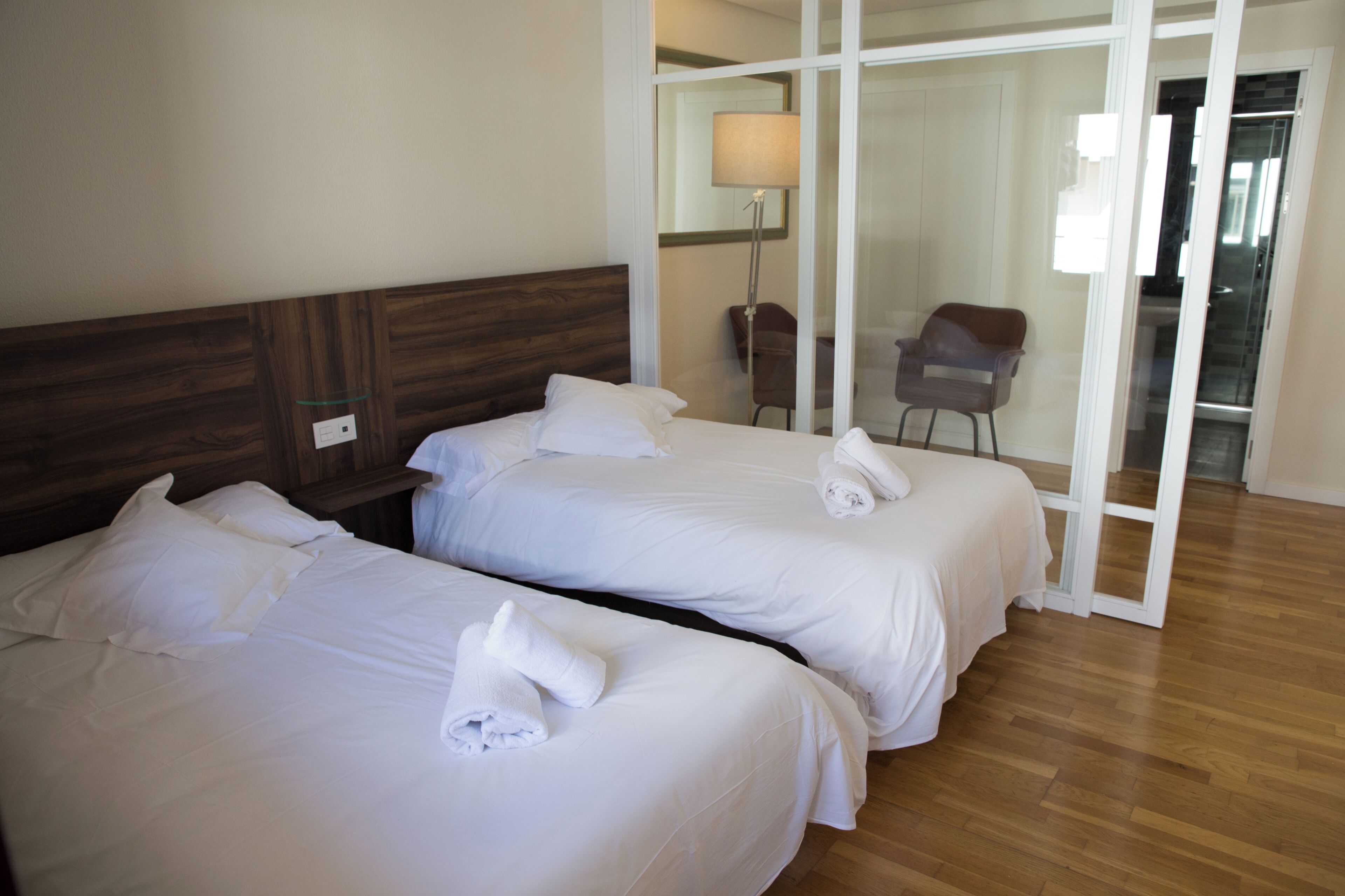 A hotel room with two beds, a wooden headboard, a chair, and a sliding glass door leading to a balcony.