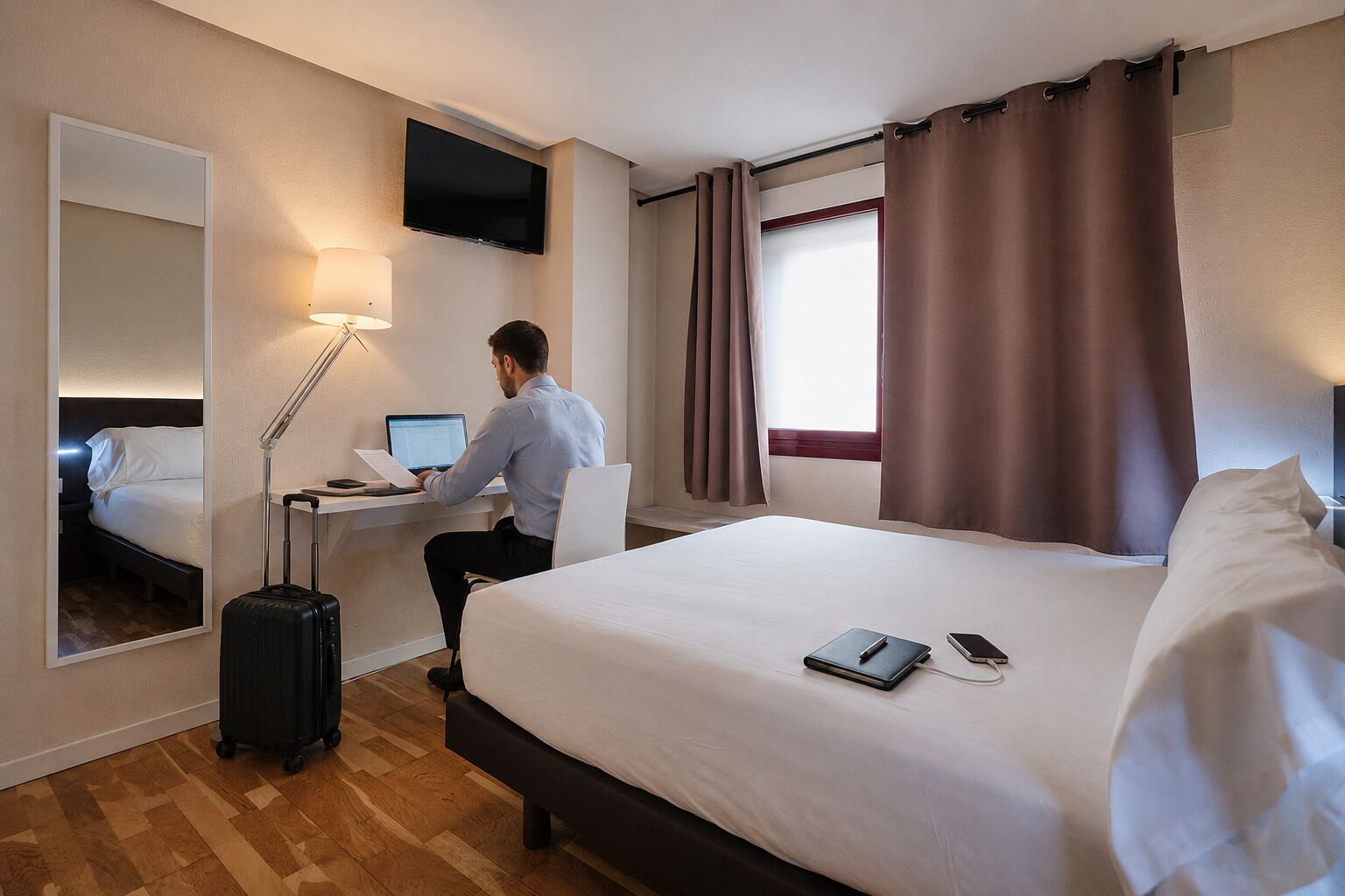 A hotel room with a bed, a desk with a laptop, a suitcase, a TV, and a window with curtains.