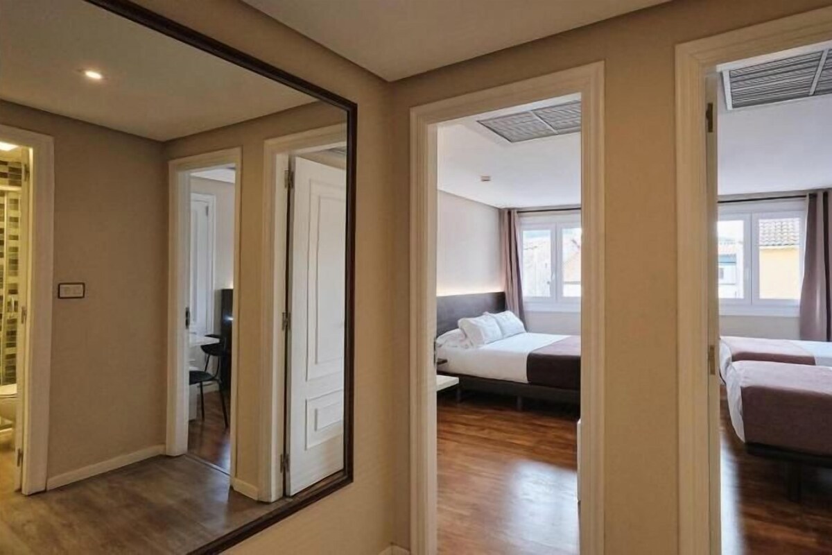 Quarto de hotel com uma cama grande, uma escrivaninha e uma cadeira. Há um espelho na parede e uma janela com cortinas.