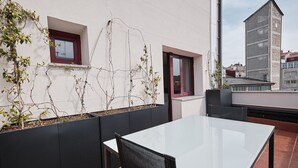 A rooftop terrace with a white table and chairs, a black planter box with plants, and a view of a cityscape. - Hotel Mogay (Chantada)