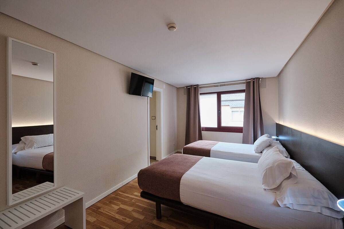 A hotel room with two beds, a television, a window with curtains, and a mirror.