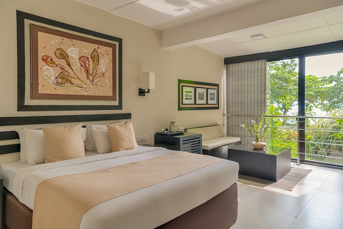 A modern hotel room with a large bed, a small desk, a balcony with a view of greenery, and artwork on the walls.