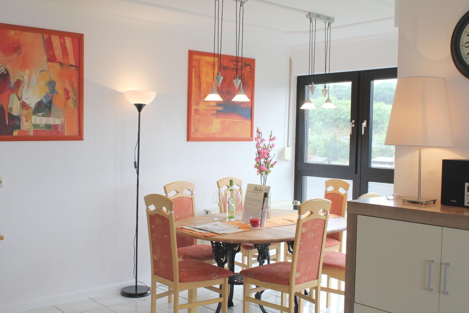 A dining area with a round table, six chairs, and hanging pendant lights. There is a window with a view of greenery outside.