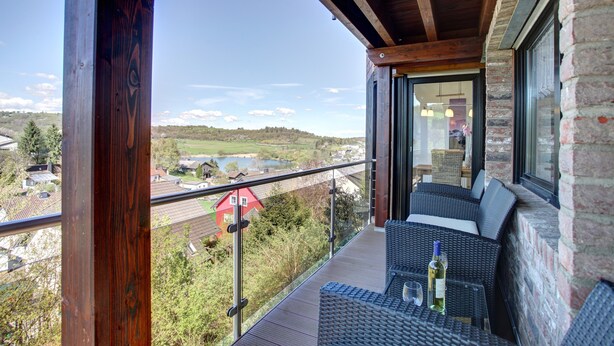 A balcony with wicker furniture, a glass table, and a bottle of wine. - Ferienwohnung Seeblick (Schalkenmehren)