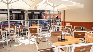 Un café en plein air avec des tables en bois et des chaises blanches, de grands parasols et une vue sur la rue grâce à des murs en verre.