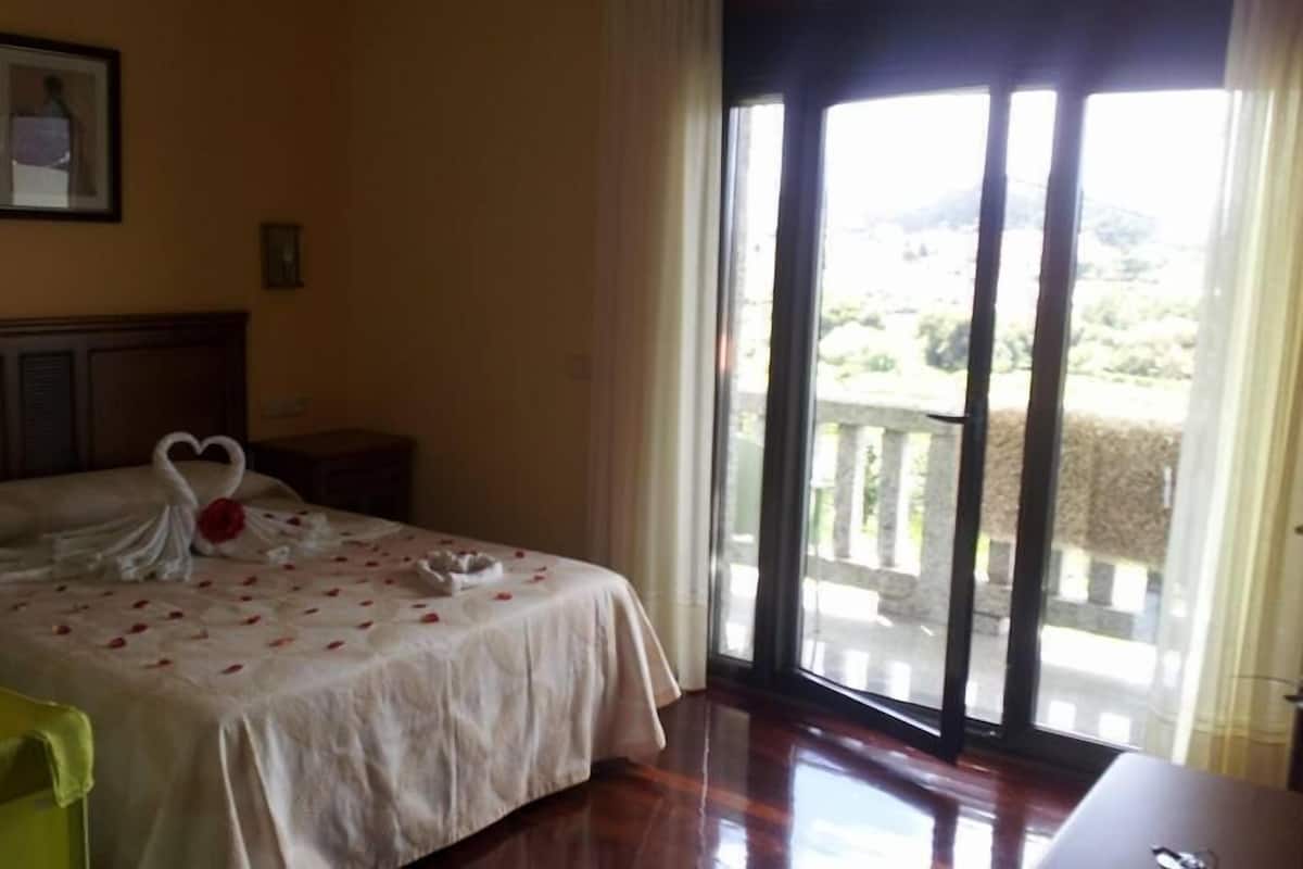 Habitación de hotel con cama, cabecera de madera, mesita de noche, una silla verde y balcón con vistas al campo.