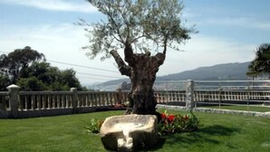 A tree with a stone base in a landscaped area with a view of a body of water and distant hills. - Hotel Liñares (Poio)