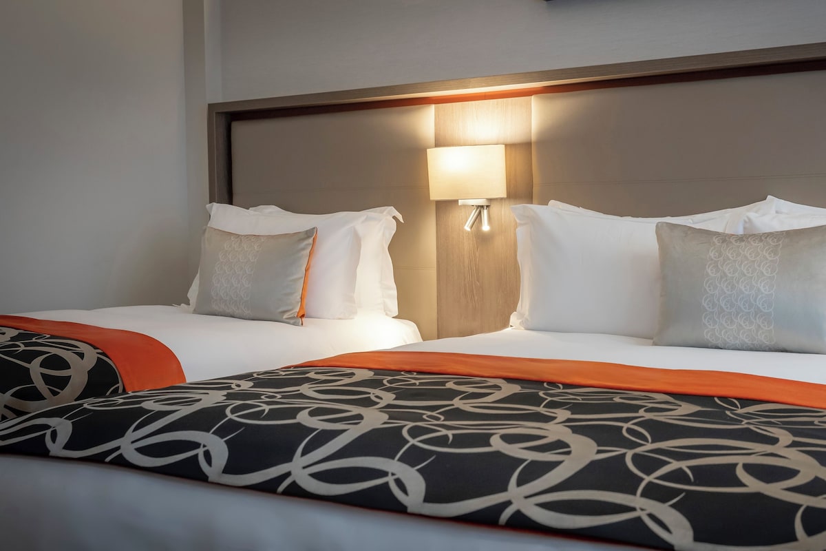 Een hotelkamer met twee bedden, elk voorzien van witte lakens en gestreepte kussens. Tussen de bedden hangt een wandlamp.
