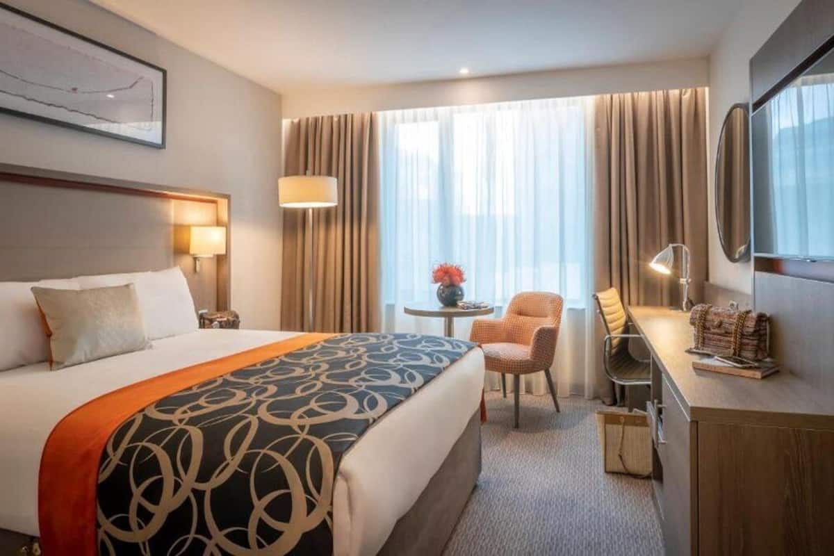 Een hotelkamer met een groot bed, een bureau, een stoel, een klein tafeltje met een vaas en een raam met gordijnen.