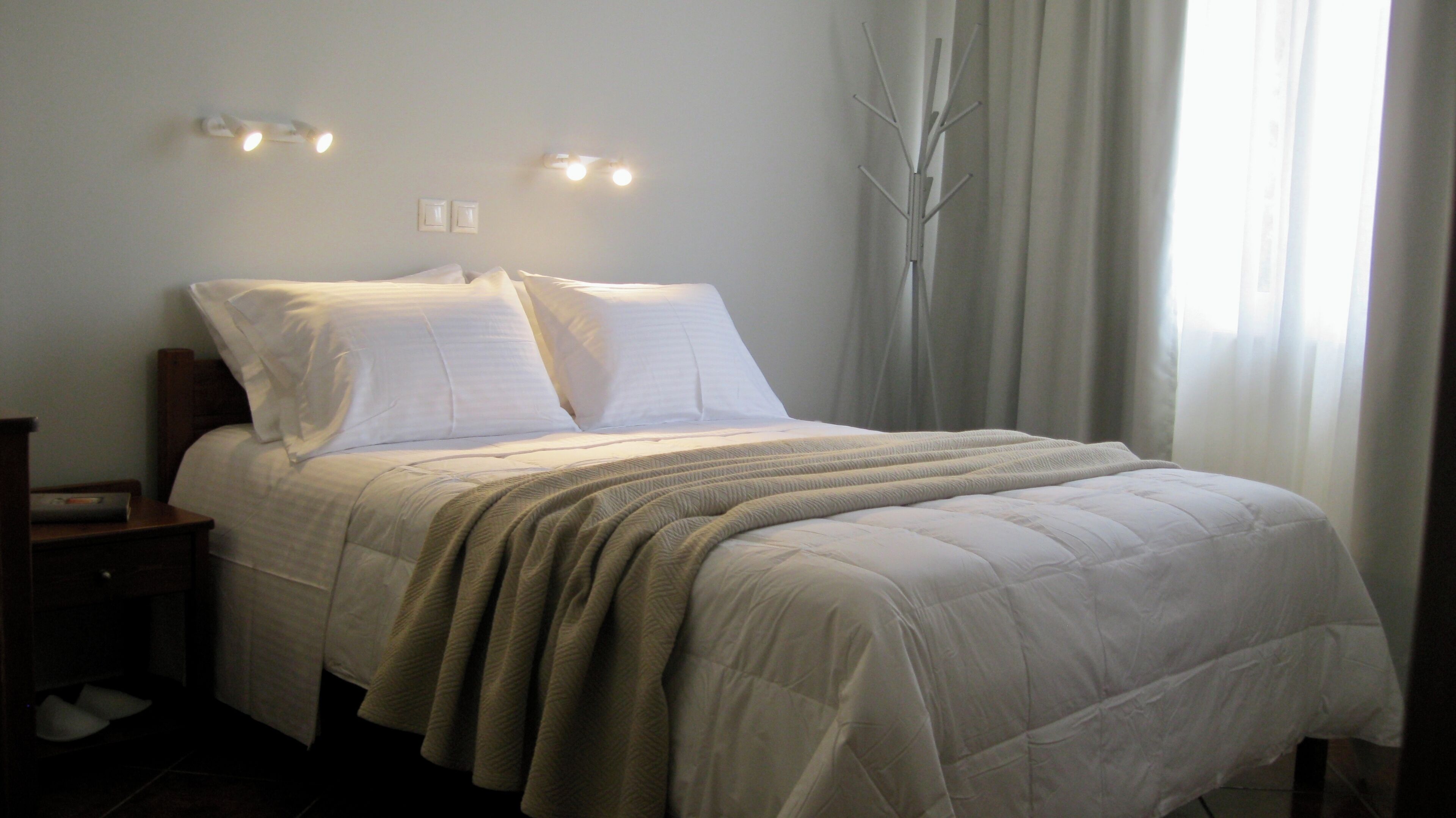 Ein ordentlich bezogenes Bett mit weißer Decke und Kissen, ein Nachttisch mit Lampe und ein Fenster mit durchsichtigen Vorhängen.